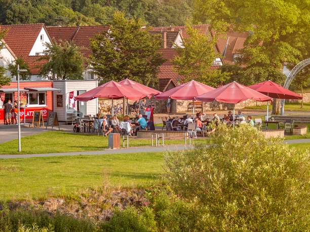 Café mit roten Sonnenschirmen im Grünen, Gäste sitzen auf der Terrasse, Kleinstadt im Hintergrund.