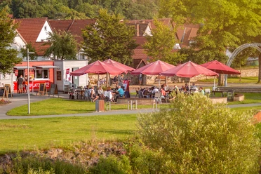 Café Ankerplatz im Emmerauenpark Lügde Café mit roten Sonnenschirmen im Grünen, Gäste sitzen auf der Terrasse, Kleinstadt im Hintergrund.