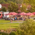 Café Ankerplatz im Emmerauenpark Lügde Café mit roten Sonnenschirmen im Grünen, Gäste sitzen auf der Terrasse, Kleinstadt im Hintergrund.