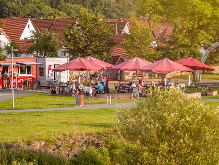Café Ankerplatz im Emmerauenpark Lügde Café mit roten Sonnenschirmen im Grünen, Gäste sitzen auf der Terrasse, Kleinstadt im Hintergrund.