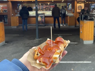 Eine Hand hält eine Currywurst auf einer Pappschale, Marktstand im Hintergrund.