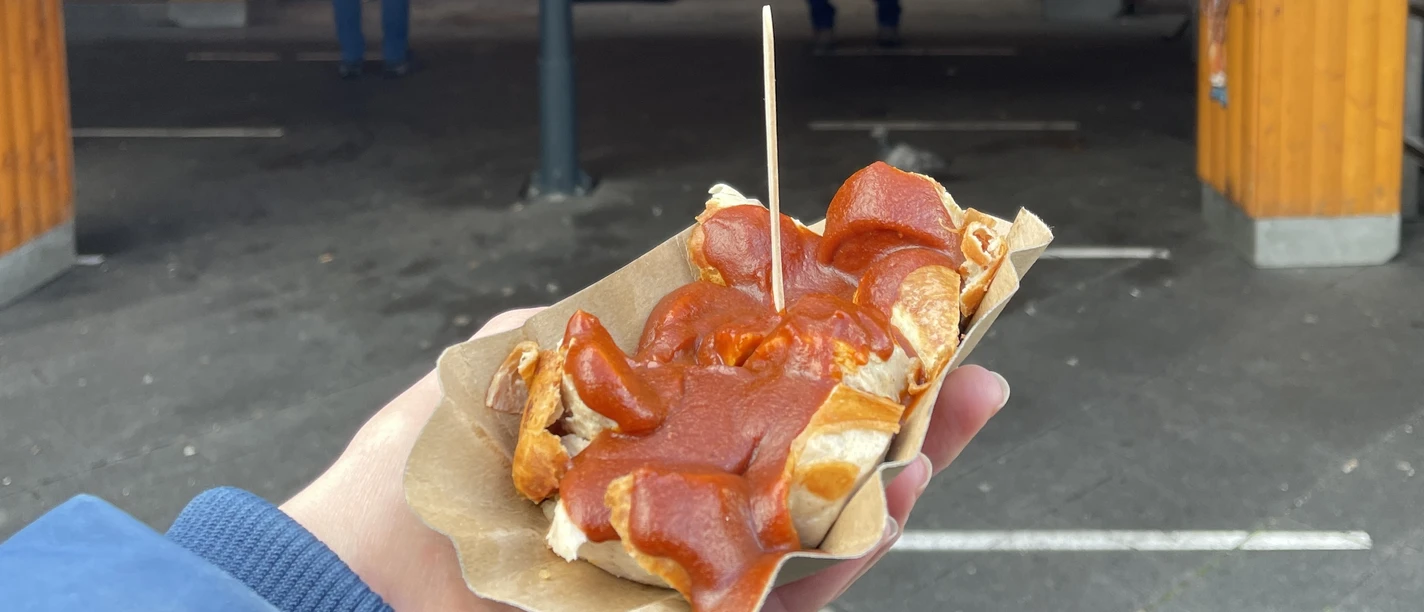 Eine Hand hält eine Currywurst auf einer Pappschale, Marktstand im Hintergrund.