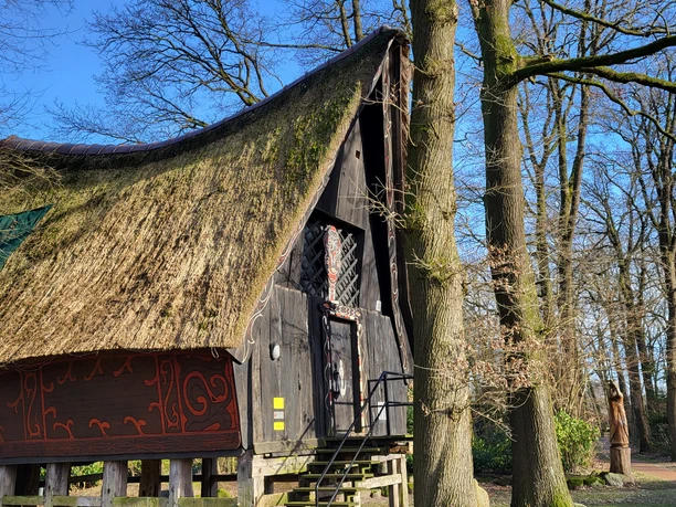 Batakhaus, Werpelo ©Naturpark Hümmling (2).jpg Pfahlhaus mit kunstvoll geschnitztem Giebel und Reetdach zwischen hohen Bäumen im Naturpark Hümmling