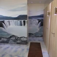 Sonnenwiese WC Öffentliches WC mit Wandgemälde eines Wasserfalls, helle Fliesen und ein Rollbahnstreifen.