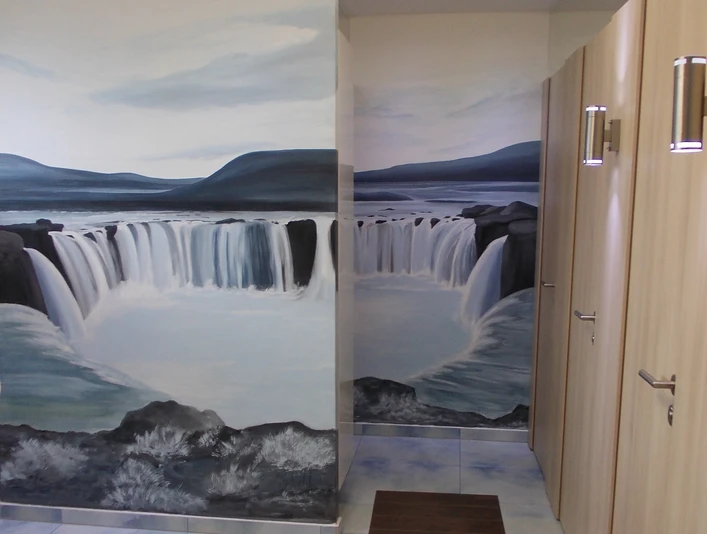 Sonnenwiese WC Öffentliches WC mit Wandgemälde eines Wasserfalls, helle Fliesen und ein Rollbahnstreifen.