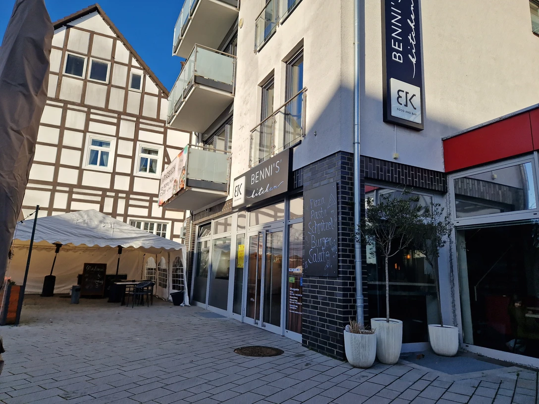 Bennis Kitchen Delbrück: Modernes Restaurant mit Glasfassade, Sonnenschirmen und Fachwerkhaushintergrund.