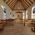 Innenansicht Hövelsenner Kapelle Hölzerne Bänke und Statuen vor lichtdurchfluteten, bunten Glasfenstern mit christlichen Motiven.