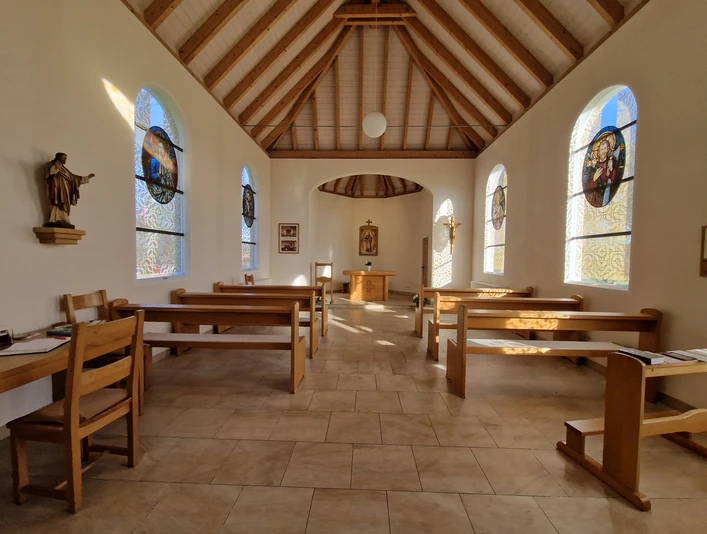 Innenansicht Hövelsenner Kapelle Hölzerne Bänke und Statuen vor lichtdurchfluteten, bunten Glasfenstern mit christlichen Motiven.