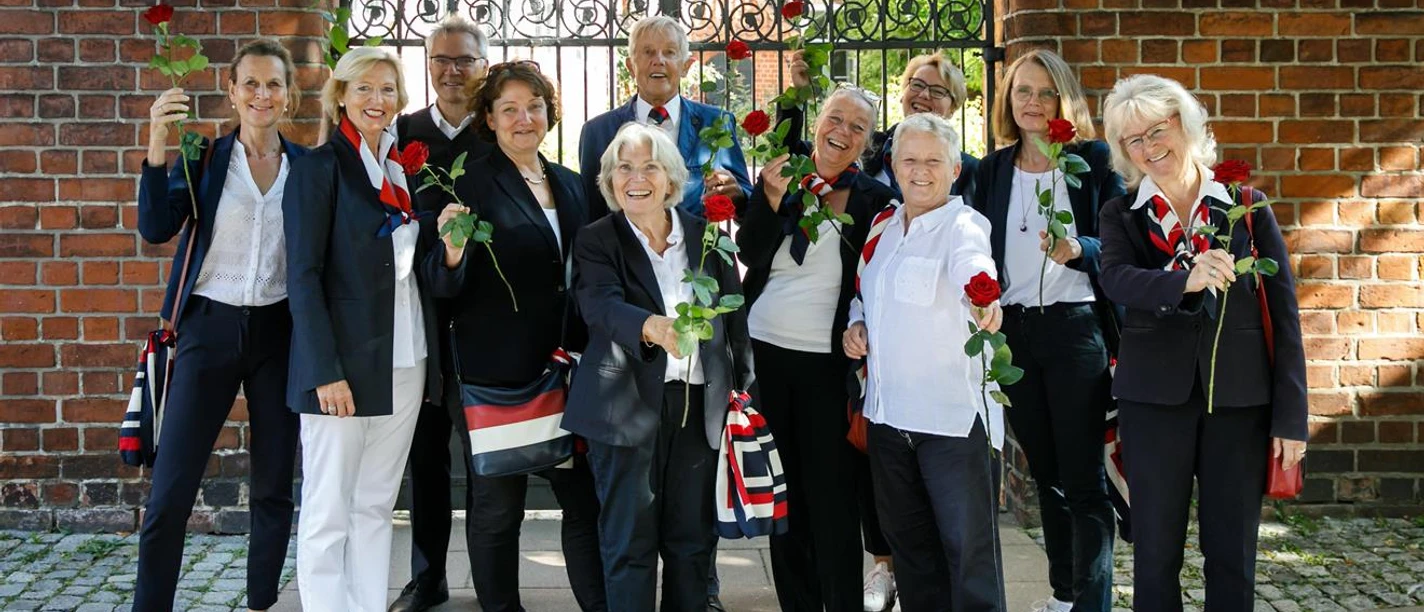 Zehn lachende Rote Rosen Stadtführer:innen halten rote Rosen vor einem Backsteintor.