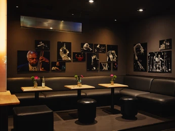 Stadtgarten Ein gemütlicher, dunkel gehaltener Raum mit schwarzen Ledersitzmöglichkeiten und Jazz-Fotografien an den Wänden.A cozy, dark room with black leather seating and jazz photographs on the walls.