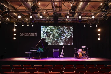 Stadtgarten Eine Bühne mit Flügel, Gitarre, Schlagzeug und "Stadtgarten"-Schild im Hintergrund.