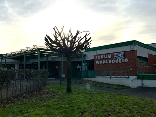 Forum Wahlscheid <p>Ansicht des Forum Wahlscheid mit einem beschnittenen Baum im Vordergrund an einem bewölkten Tag.</p>