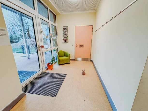 Kindergarten-Rehlingen-2024 (1).jpg Heller Eingangsbereich mit Glastür, grünem Sessel und rosa Küchentür.