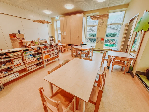 Kindergarten-Rehlingen-2024 (10).jpg Heller Gruppenraum im Kindergarten Rehlingen mit Holztischen, Spielregalen und großen Fenstern.