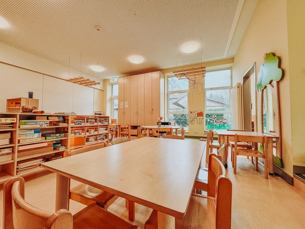 Kindergarten-Rehlingen-2024 (11).jpg Heller Kindergartenraum mit Holztischen, Stühlen und Regalen voller Spiele.