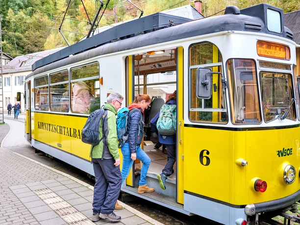 People board a yellow Kirnitzschtalbahn streetcar, surrounded by green countryside and buildings in the background.