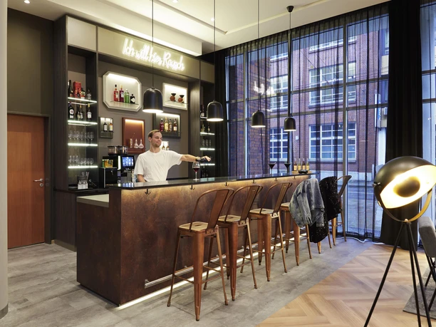 Eine moderne Bar mit Holzregalen und einer eleganten Theke. Im Hintergrund steht ein Barkeeper, der Getränke serviert, während große Fenster Tageslicht hereinlassen.