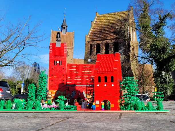 bricks-am-meer-kirche.jpg Rote Backsteinkirche mit passendem LEGO-Modell im Vordergrund, umgeben von Bäumen und Autos.