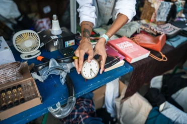 Odonien A person holds an old alarm clock above a flea market stall with various everyday objects.