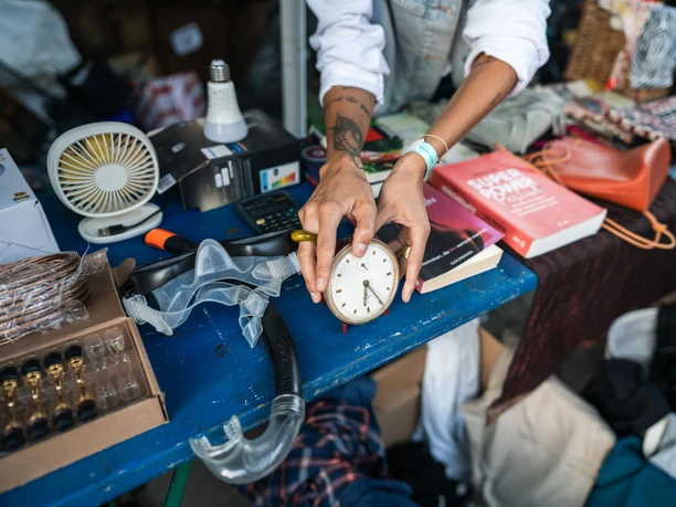 Odonien A person holds an old alarm clock above a flea market stall with various everyday objects.