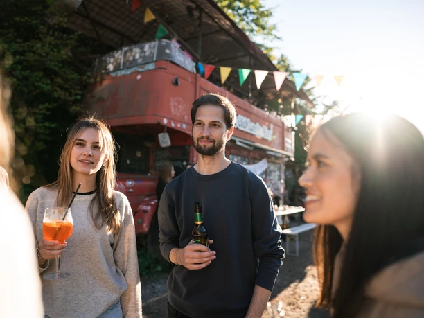 Odonien Eine Gruppe junger Menschen steht vor einem roten Doppeldeckerbus in einer entspannten, urbanen Atmosphäre.