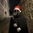 Eine Figur im mittelalterlichen Pestdoktor-Outfit mit Weihnachtsmütze steht in einer dunklen Gasse.