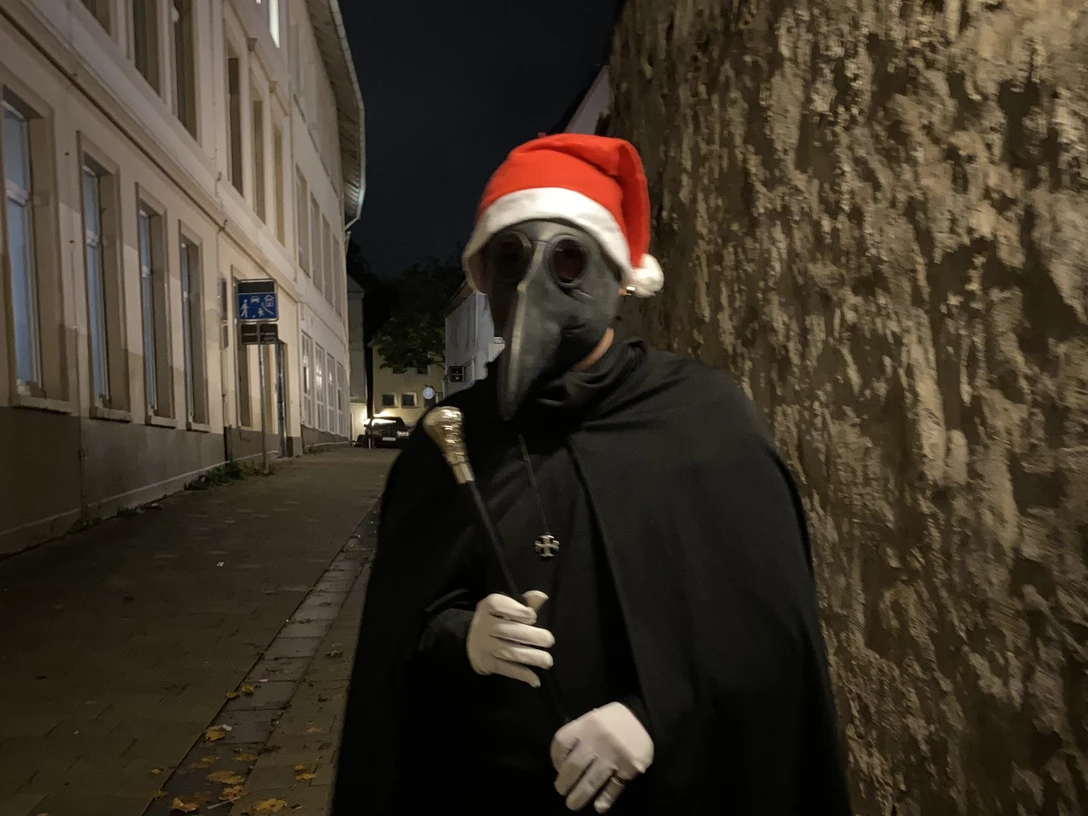 IMG_0388.jpg2.jpg Eine Figur im mittelalterlichen Pestdoktor-Outfit mit Weihnachtsmütze steht in einer dunklen Gasse.