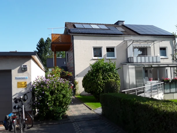 Hausansicht mit zwei Balkonen an der Fronseite, seitlich im oberen Stockwerk ein weiterer Balkon, Solarmodule auf dem Dach, im vorderen Bereich eine grüne Hecke und Büsche