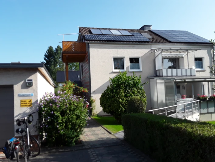 Hausansicht mit zwei Balkonen an der Fronseite, seitlich im oberen Stockwerk ein weiterer Balkon, Solarmodule auf dem Dach, im vorderen Bereich eine grüne Hecke und Büsche
