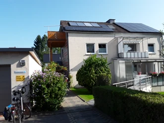 Haus Rasche Hausansicht mit zwei Balkonen an der Fronseite, seitlich im oberen Stockwerk ein weiterer Balkon, Solarmodule auf dem Dach, im vorderen Bereich eine grüne Hecke und Büsche