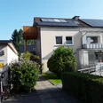 Haus Rasche Hausansicht mit zwei Balkonen an der Fronseite, seitlich im oberen Stockwerk ein weiterer Balkon, Solarmodule auf dem Dach, im vorderen Bereich eine grüne Hecke und Büsche