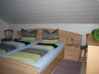 Schlafzimmer mit Doppelbett aus Holz, zwei Nachtschränken mit jeweils einer Leselampe, links und rechts vom Bett ein Holzstuhl, rechts eine Kommode