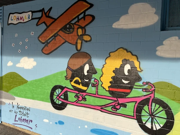 Wandgestaltung an der Jabachhalle Wandgemälde in Lohmar zeigt Comicfiguren auf einem Tandem, über ihnen fliegt ein Flugzeug.