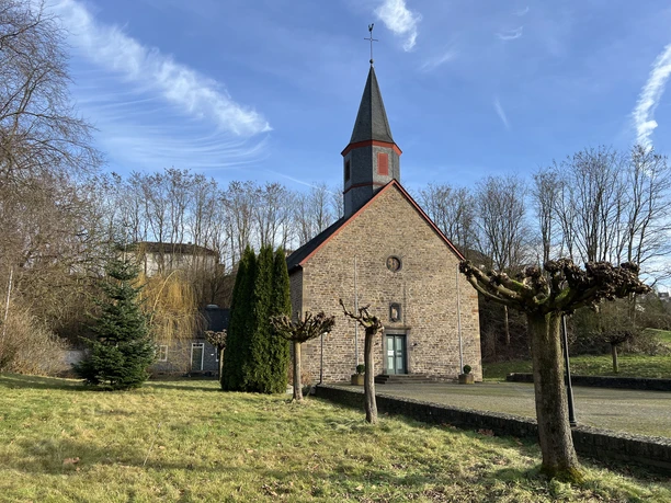 kath. Kirche Wahlscheid <p>Stein-Backsteinkirche mit rotem Dach und Kirchturm, umgeben von Bäumen und grasbewachsener Wiese.</p>