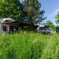 Auto mit zelt.jpg Auto und Zelt stehen inmitten einer grünen Wiese, umgeben von Bäumen, unter blauem Himmel.