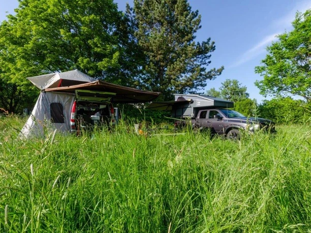 Auto mit zelt.jpg Auto und Zelt stehen inmitten einer grünen Wiese, umgeben von Bäumen, unter blauem Himmel.