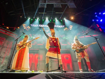 Stunksitzung Drei kostümierte Personen stehen auf einer Bühne, beleuchtet von buntem Scheinwerferlicht.Three costumed people stand on a stage, illuminated by colorful spotlights.