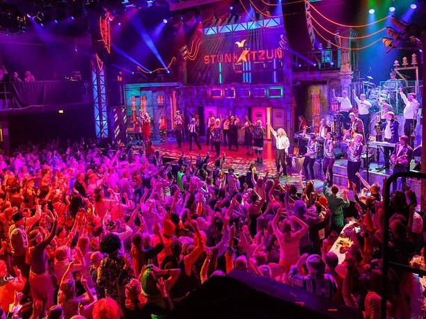 Stunksitzung A lively scene from the Cologne Stunksitzung. A stage is occupied by a colorful cabaret group, while the enthusiastic audience applauds exuberantly and fills the festive atmosphere.