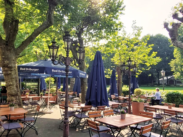 Geräumiger Biergarten mit belaubten Bäumen, Holztischen, blauen Sonnenschirmen und ruhiger Atmosphäre.
