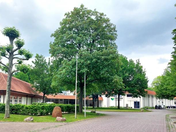 Hotelgebäude mit roten Ziegeldächern, umgeben von gepflegten Bäumen und einem gepflasterten Weg.