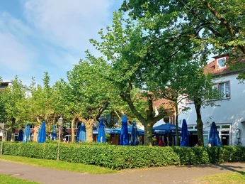Biergarten des Schützenhofs Paderborn Tische, Stühle und Sonnenschirme unter Bäumen in einem Biergarten neben einem weißem Gebäude.