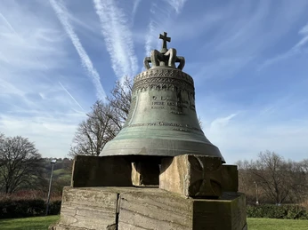 Glocke vor der ev. Kirche Die Gedenk-Glocke in Lohmar, montiert auf einem Holzsockel, vor blauem Himmel und Bäumen.