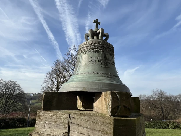 Glocke vor der ev. Kirche Die Gedenk-Glocke in Lohmar, montiert auf einem Holzsockel, vor blauem Himmel und Bäumen.