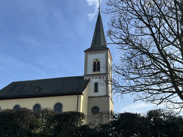ev. Bartholomäuskirche Die Kirche in Lohmar mit spitzem Turm und umliegenden Bäumen vor blauem Himmel.