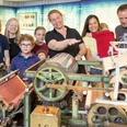 Familie an der Laborpapiermaschine Eine Familie erforscht eine historische Papiermaschine in einem Museum, geführt von einem Mitarbeiter.