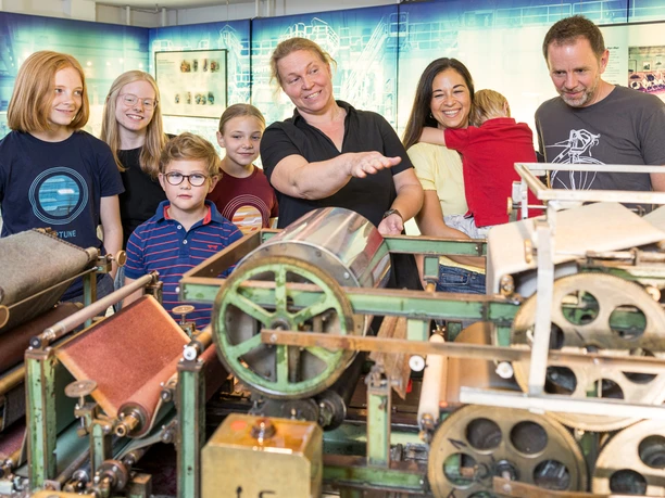 Familie an der Laborpapiermaschine Eine Familie erforscht eine historische Papiermaschine in einem Museum, geführt von einem Mitarbeiter.