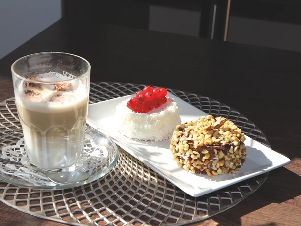 Ein Latte Macchiato neben zwei kunstvoll dekorierten Törtchen mit Beeren und Nüssen auf einem Tisch.