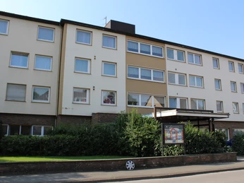 Vierstöckiges Wohngebäude mit beige-weißer Fassade, zahlreichen Fenstern und einem überdachten Eingang.