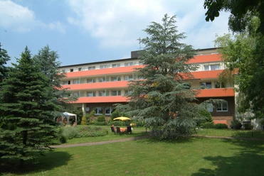 Vierstöckiges Gebäude mit oranger Fassade, umgeben von gepflegtem Garten und grünen Bäumen.
