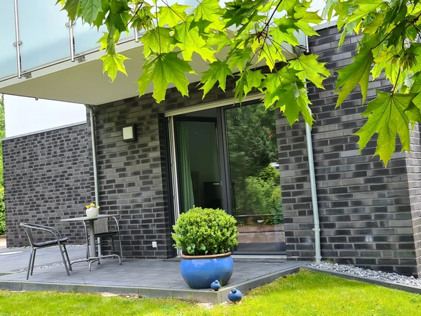 FeWo am Park Außenansicht.jpg Moderne Ferienwohnung mit dunkler Klinkerfassade, Terrasse und Gartenmöbeln in grüner Umgebung.